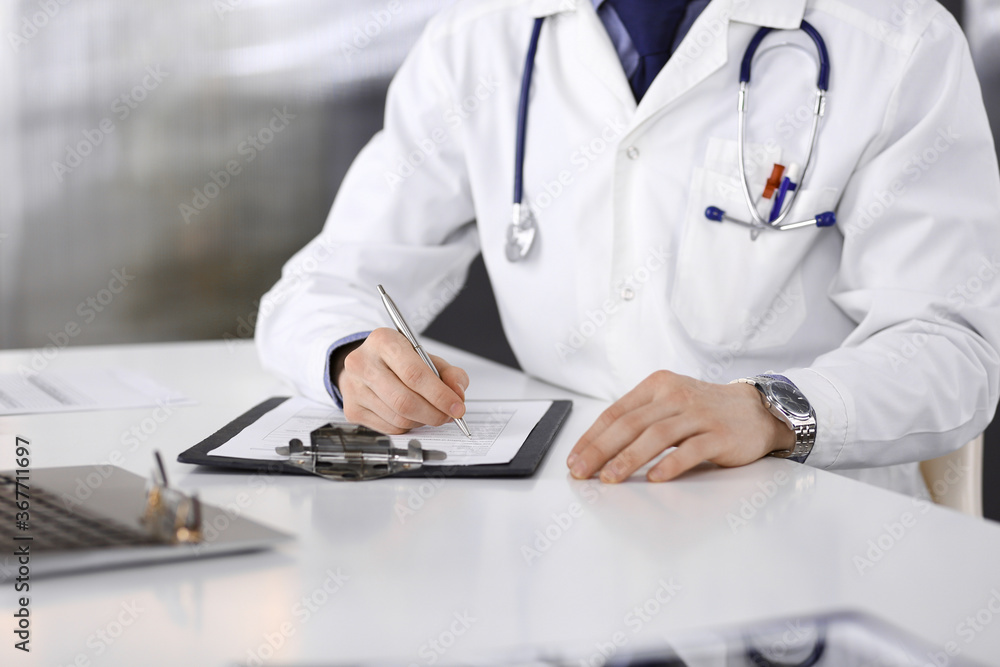 Unknown male doctor sitting and working with clipboard of medication history record in clinic at his working place, close-up. Young physician at work. Perfect medical service, medicine concept Unknown male doctor sitting and working with clipboard of medication history record in clinic at his working place, close-up. Young physician at work. Perfect medical service, medicine concept