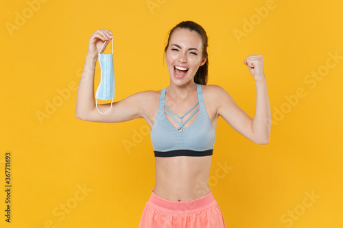 Happy young fitness sporty woman girl in sportswear working out isolated on yellow wall background. Workout sport motivation concept. Mock up copy space. Hold sterile face mask, doing winner gesture.