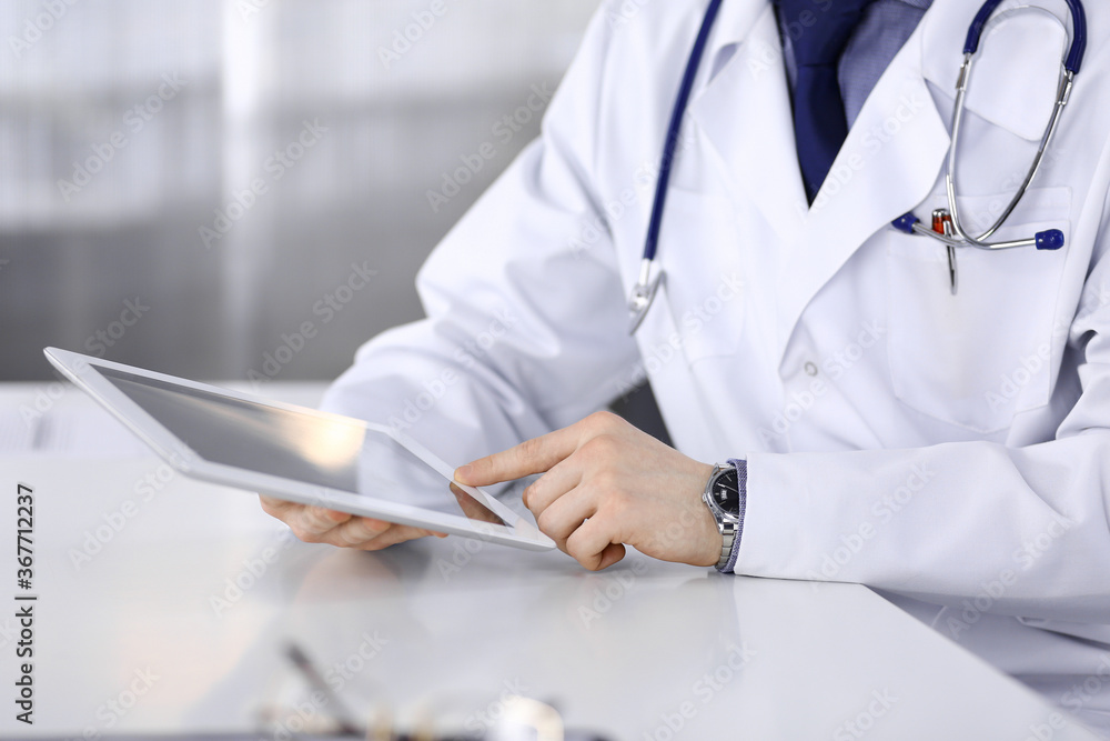 Unknown male doctor sitting and working with tablet computer in clinic at his working place, close-up. Young physician at work. Perfect medical service, medicine concept Unknown male doctor sitting and working with tablet computer in clinic at his working place, close-up. Young physician at work. Perfect medical service, medicine concept
