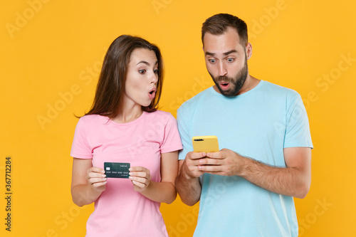 Shocked young couple two friends guy girl in blue pink t-shirts isolated on yellow background studio portrait. People lifestyle concept. Mock up copy space. Using mobile phone, hold credit bank card. © ViDi Studio