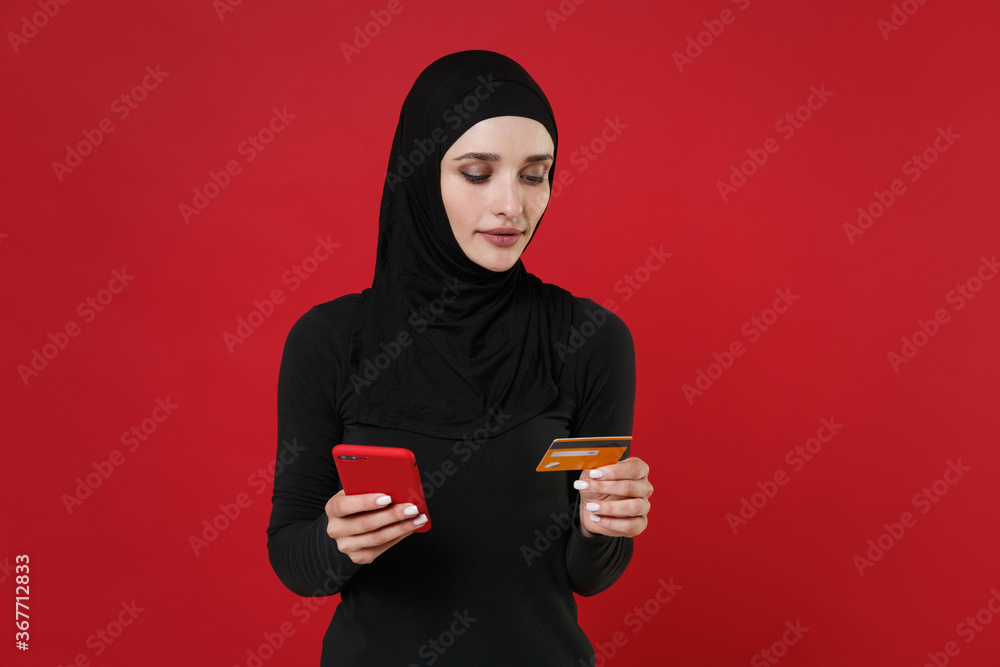 Beautiful young arabian muslim woman in hijab black clothes posing isolated on red background studio. People religious lifestyle concept. Mock up copy space. Using mobile phone, hold credit bank card. Beautiful young arabian muslim woman in hijab black clothes posing isolated on red background studio. People religious lifestyle concept. Mock up copy space. Using mobile phone, hold credit bank card.