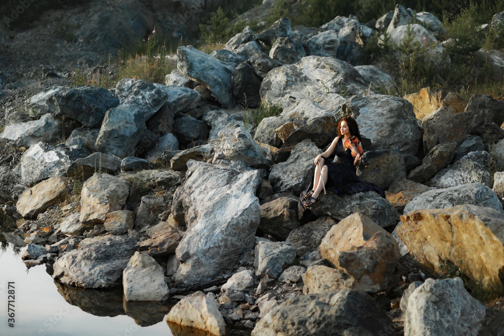 brunette girl with lush curly hair sits on the stones near the lake in summer in a dark blue dress with a violin, professional musician violinist