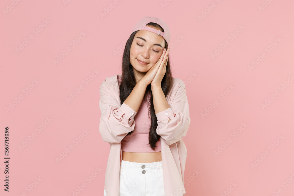 Smiling young asian woman girl in casual clothes cap posing isolated on pastel pink wall background studio portrait. People lifestyle concept. Mock up copy space. Sleep with folded hands under cheek.