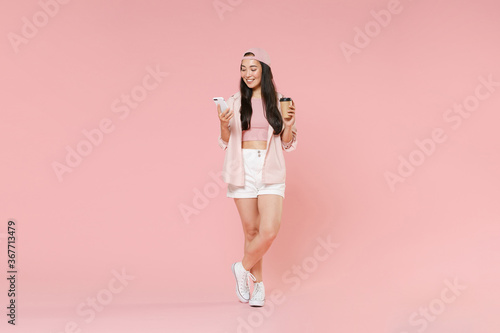 Full length portrait of smiling asian girl in casual clothes, cap isolated on pink background. People lifestyle concept. Mock up copy space. Using mobile cell phone, hold paper cup of coffee or tea.
