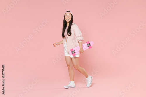 Full length portrait of smiling young asian girl in casual clothes, cap isolated on pastel pink background studio portrait. People lifestyle concept. Mock up copy space. Hold skateboard looking aside.