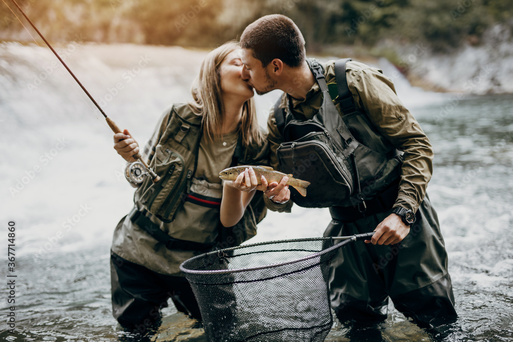 Young adult couple is fishing together on fast mountain river. They holds a live trout and kisses each other before releasing it into the river again. Young adult couple is fishing together on fast mountain river. They holds a live trout and kisses each other before releasing it into the river again.