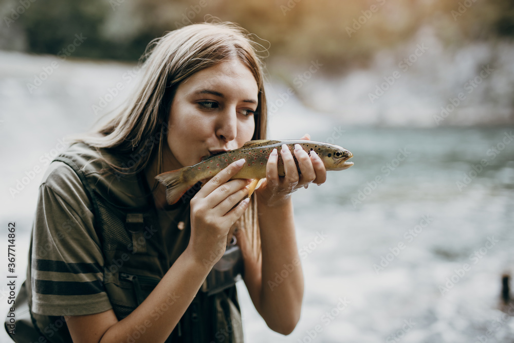 Young adult woman is fishing alone on fast mountain river. The girl holds a  live trout and kisses it before releasing it into the river again. Stock  Photo
