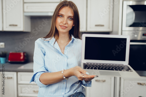 Young blonde woman standing in kithen and showing blank screen laptop computer
