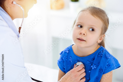 Doctor examining a child by stethoscope in sunny clinic. Happy smiling girl patient dressed in blue dress is at usual medical inspection