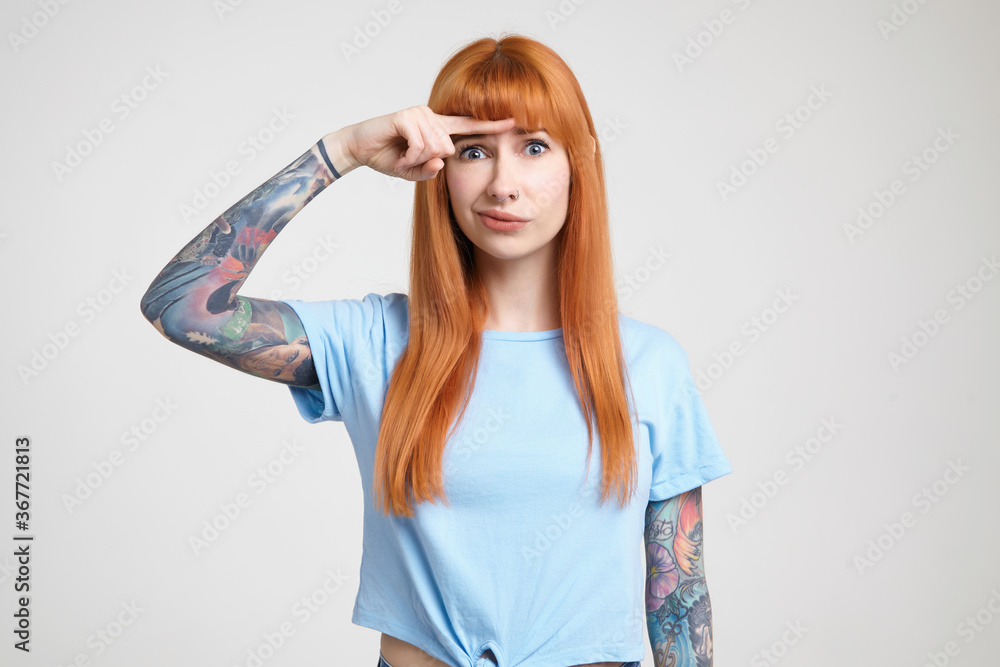 Indoor photo of young long haired redhead woman with tattoos keeping forefinger above her eyebrows and grimacing her face while looking at camera, isolated over white background Indoor photo of young long haired redhead woman with tattoos keeping forefinger above her eyebrows and grimacing her face while looking at camera, isolated over white background
