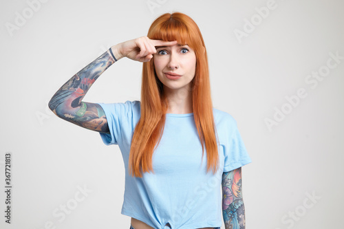Indoor photo of young long haired redhead woman with tattoos keeping forefinger above her eyebrows and grimacing her face while looking at camera, isolated over white background