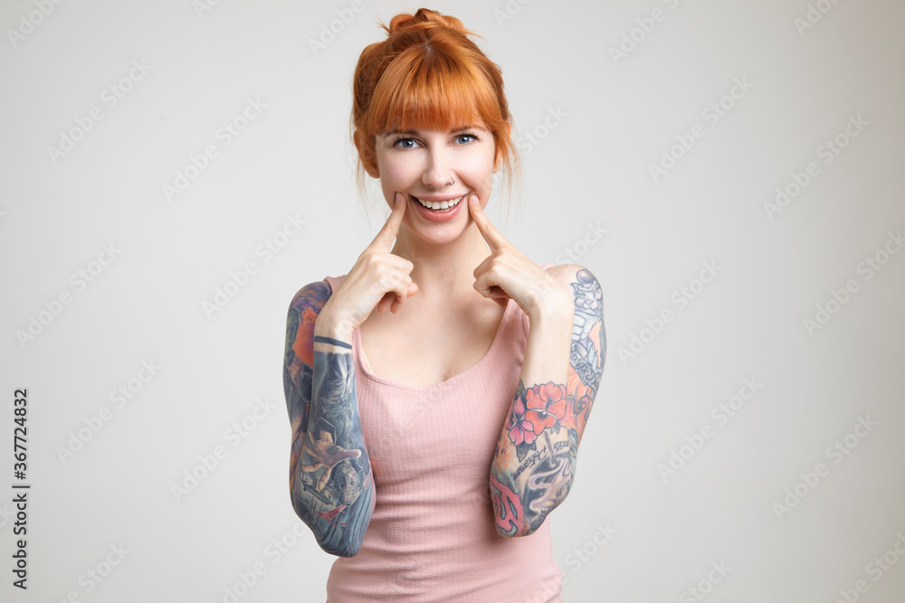 Joyful green-eyed young redhead woman with tattoos holding index fingers on her cheeks while smiling widely at camera, isolated against white background Joyful green-eyed young redhead woman with tattoos holding index fingers on her cheeks while smiling widely at camera, isolated against white background