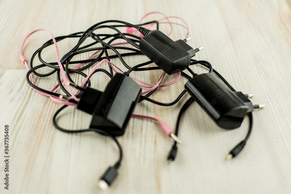 the need for universal battery charger: black 3 different chargers with different connectors and earphones tangled cables together, on the white oak table, in short focus the need for universal battery charger: black 3 different chargers with different connectors and earphones tangled cables together, on the white oak table, in short focus