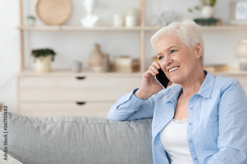 Smiling senior lady talking on cellphone while relaxing on couch at home
