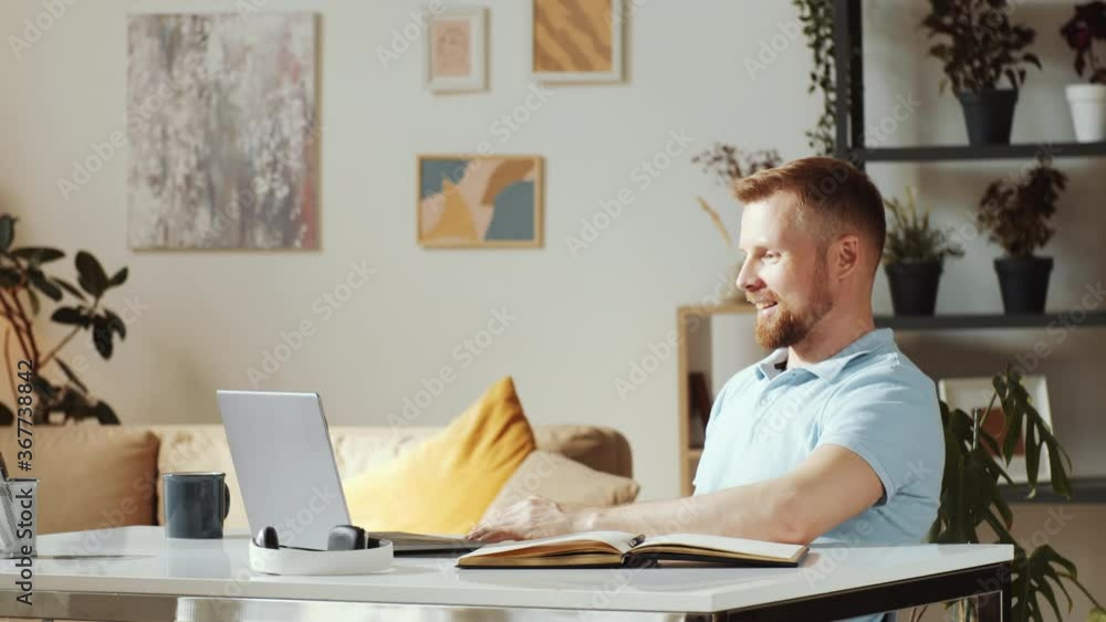 Young man smiling at waving at web camera while video calling on laptop at table in the living room Young man smiling at waving at web camera while video calling on laptop at table in the living room