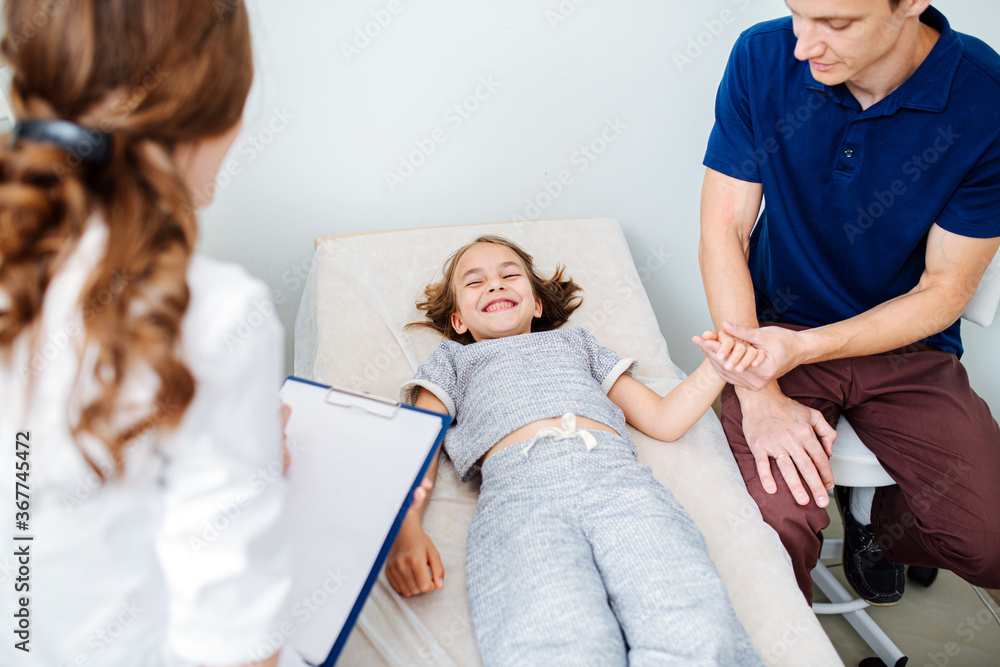 Father came with daughter to female doctor for ultrasound examination. Little girl lying on a couch, attentive father holding her hand. Doctor filling in patient's history.