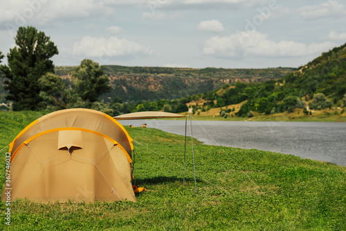 Camping beige tent in nature with view at river and mountains with green trees. Rest with a tent by the river. The pleasure of nature. Fresh air. Copy space.