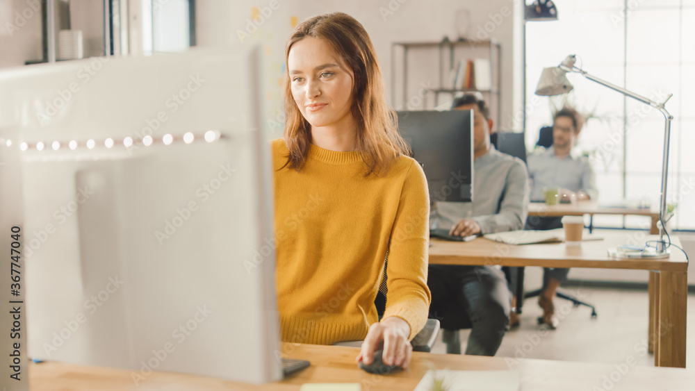 Beautiful and Smart Red Haired Female Specialist Sitting at Her Desk Works on a Desktop Computer. In the Background Modern Bright Office with Diverse Group of Professionals Working for Growing Startup Beautiful and Smart Red Haired Female Specialist Sitting at Her Desk Works on a Desktop Computer. In the Background Modern Bright Office with Diverse Group of Professionals Working for Growing Startup
