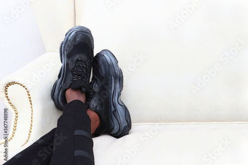 Men's tanned legs in black fashionable sneakers lie on a light sofa . An unrecognizable photo. Just my legs. Copy of the space. Light background.