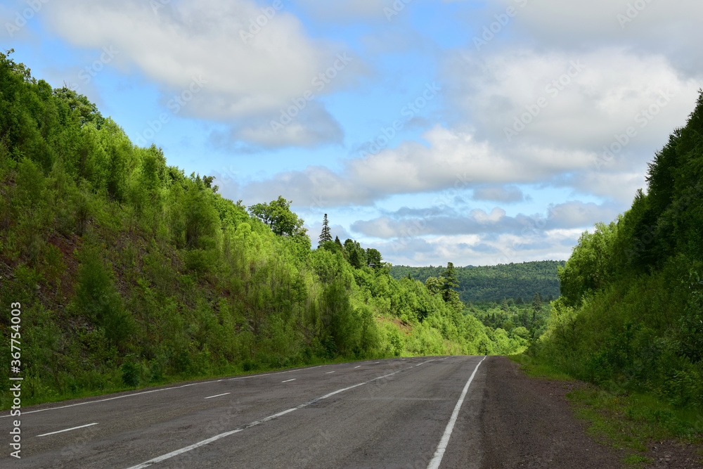Fototapeta premium Paved road among green hills. Blue sky with white clouds. Bright summer day.