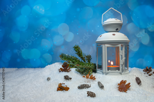 White Christmas lantern on snow with pine cones, small cones, leaves and a fir branch in front of dark blue background