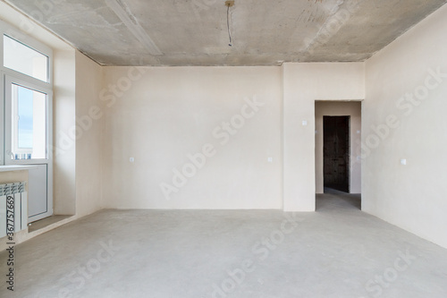 The front view an unfinished residential apartment with the white plastered walls  the empty doorway and the plastic balcony door
