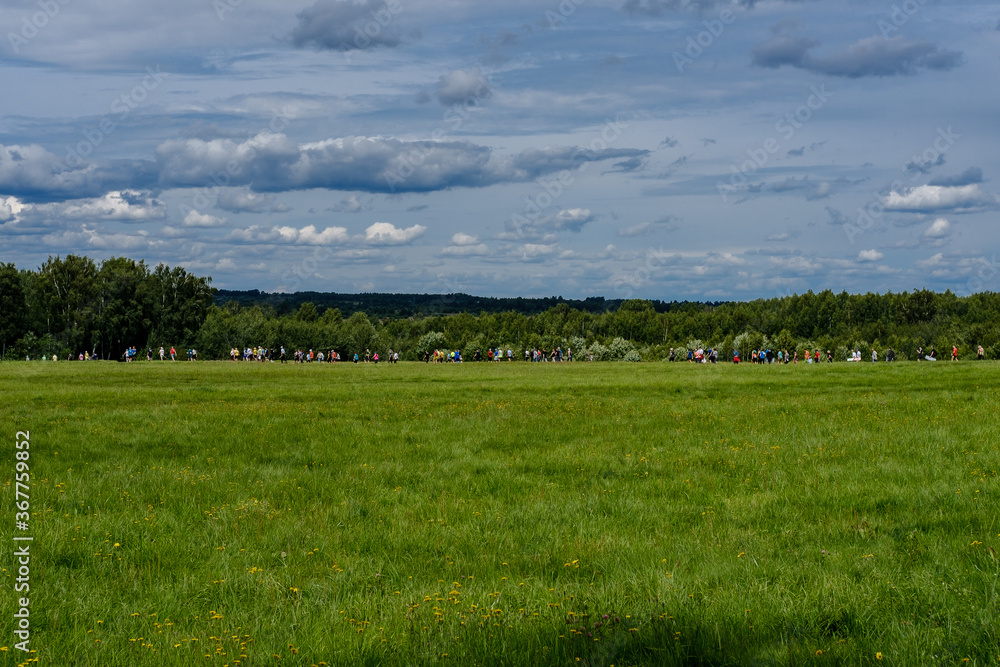 Many tourists in colorful clothes are walking through a field with green grass, and in the background a dense forest and a sky with clouds can be seen.