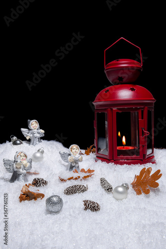 Red Christmas lantern on snow with small cones, leaves, silver choralling angles in front of black background, copy space