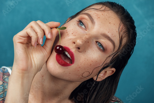 Close-up portrait of young freckled woman that try to bite cherry berry. © Grigoriy