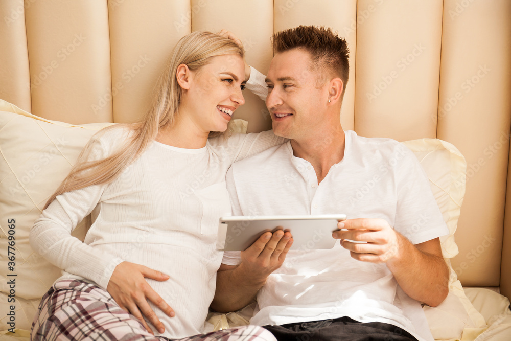 pregnant woman and a man are lying on the bed with tablet