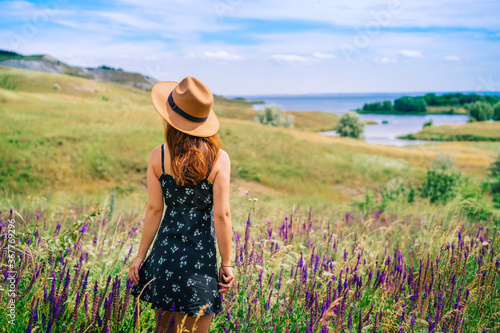 A happy young woman in a dress and hat stands against a background of blooming wildflowers and tall grass in a hilly area