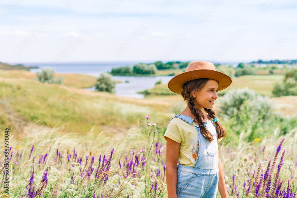 Portrait of a charming little girl in her mother's hat holding apples in her hand against a picturesque hilly landscape with a lake