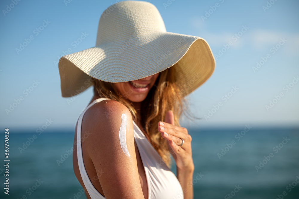 A happy smiling young woman with straw hat and white bikini is applying a sunscreen or sun tanning lotion on a shoulder to take care of her skin on a seaside beach during holidays vacation.