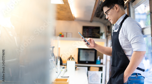 Portrait of confident Asian female barista standing inside bus coffee café. SME Business owner use smart phone for online marketing.