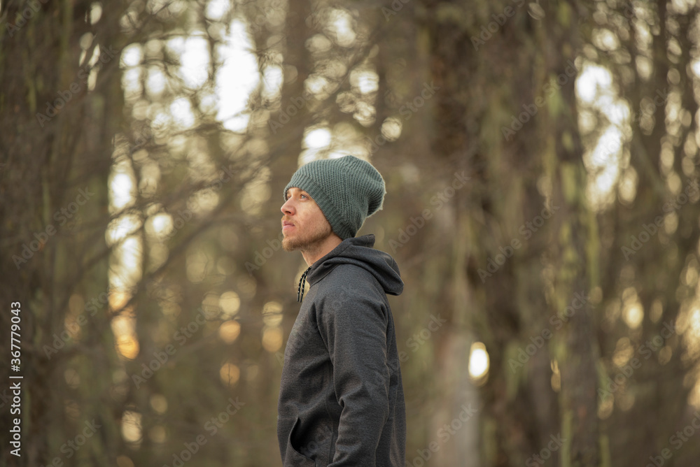Side view of a centralized man in the forest wearing gray hoddie and beanie with a winter forest as background Side view of a centralized man in the forest wearing gray hoddie and beanie with a winter forest as background