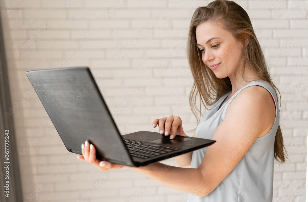 Young beautiful woman standing and using a laptop, dressed in a blue bllouse on background brick wall. Young beautiful woman standing and using a laptop, dressed in a blue bllouse on background brick wall.