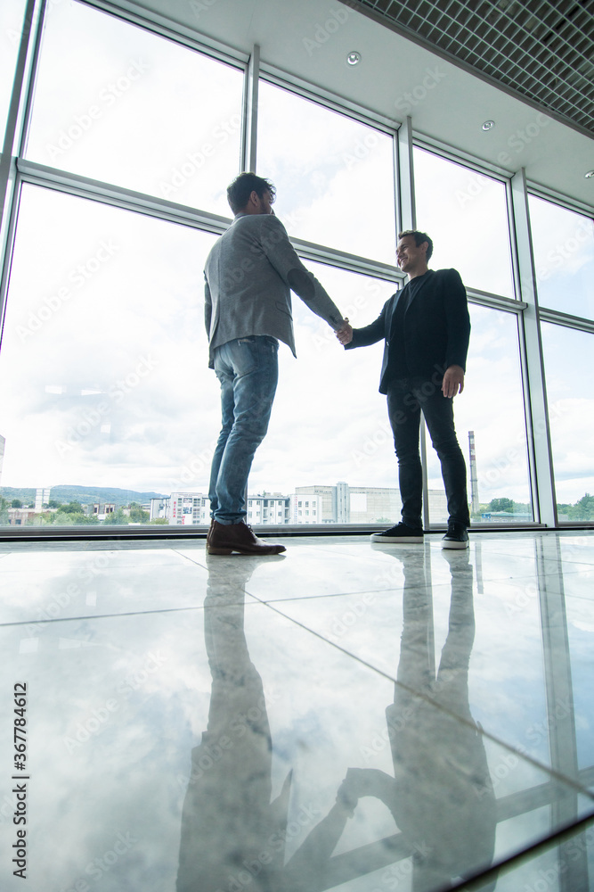 Fototapeta premium Full length side view of businessmen shaking hands in office building windows background