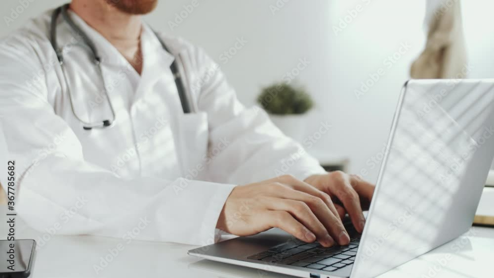 Midsection shot of male doctor in lab coat with stethoscope over his neck typing on laptop while working at desk in medical office Midsection shot of male doctor in lab coat with stethoscope over his neck typing on laptop while working at desk in medical office
