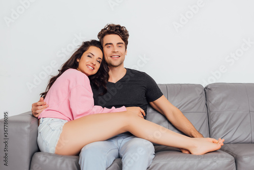 Young man hugging girlfriend and smiling at camera on couch isolated on grey