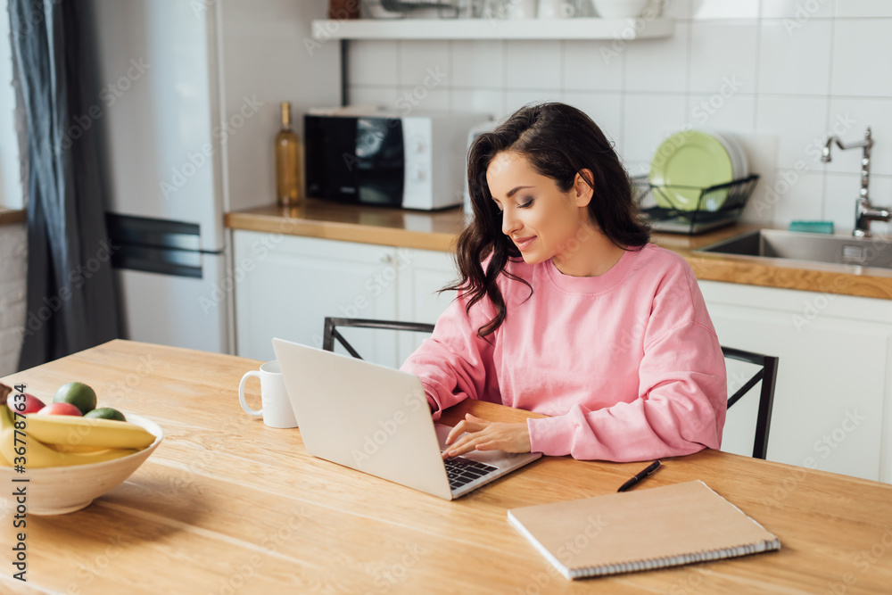 Selective focus of beautiful freelancer using laptop near notebook and cup of coffee in kitchen Selective focus of beautiful freelancer using laptop near notebook and cup of coffee in kitchen