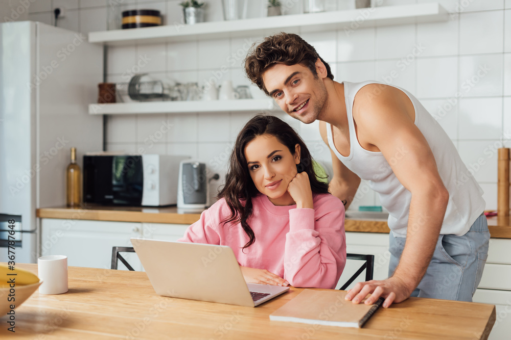 Selective focus of smiling couple looking at camera near laptop, notebook and cup of coffee on kitchen table Selective focus of smiling couple looking at camera near laptop, notebook and cup of coffee on kitchen table