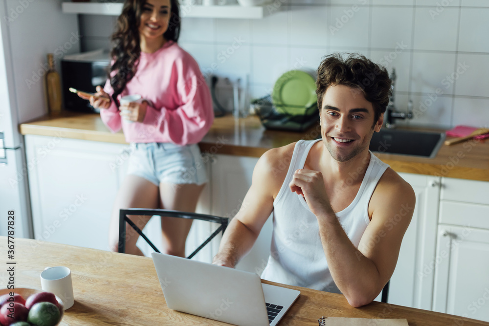 Selective focus of smiling man using laptop near girlfriend with cup and smartphone in kitchen Selective focus of smiling man using laptop near girlfriend with cup and smartphone in kitchen
