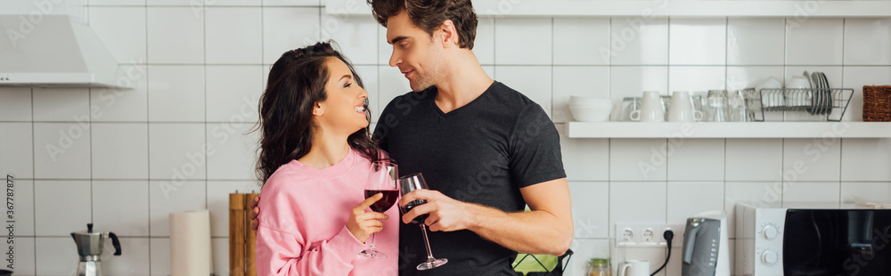 Panoramic shot of smiling woman looking at boyfriend while toasting with wine in kitchen Panoramic shot of smiling woman looking at boyfriend while toasting with wine in kitchen