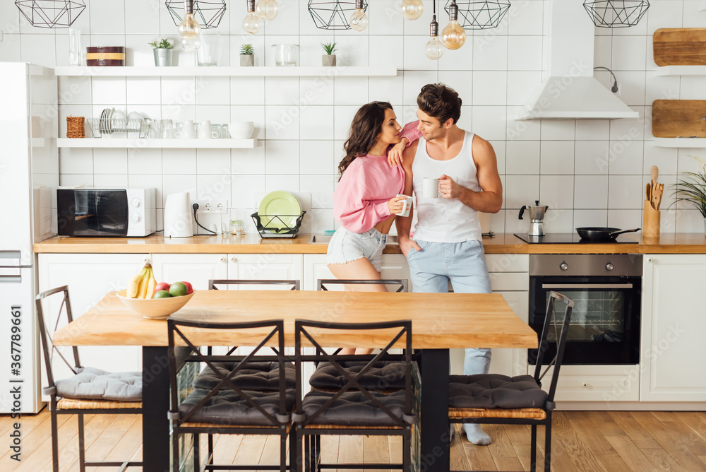 Selective focus of sexy woman embracing boyfriend with cup of coffee in kitchen Selective focus of sexy woman embracing boyfriend with cup of coffee in kitchen