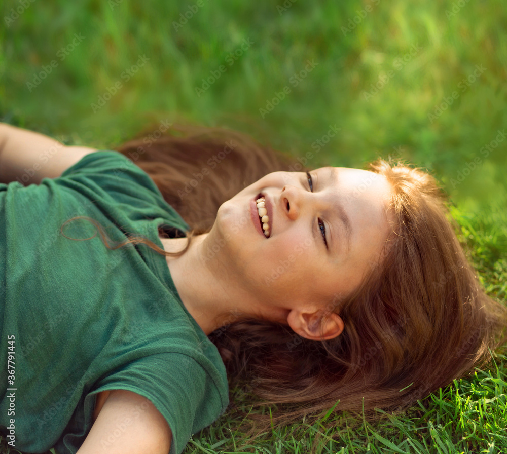 Laughing playful kid girl lying on the grass on nature summer background and looking up. Closeup positive outdoors