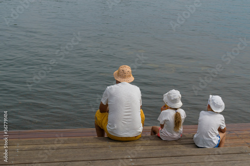 A man and children are sitting on a wooden pier by the sea. View from the back. White T-shirt, Panama hat. Concept - family vacation, father and children.