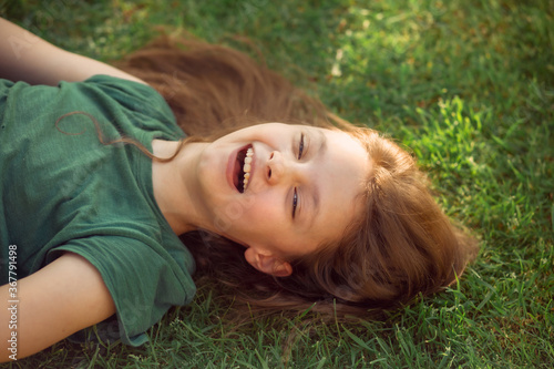 Happy laughing fun kid girl lying on the grass on nature summer background. Closeup positive outdoors bright sunny
