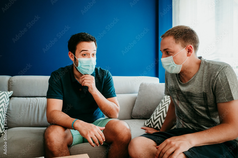 .Two friends chatting together on the sofa at home. They wear a surgical mask to take care of the covid 19 during the pandemic. Social distance