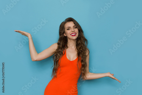 Cheerful Young Woman In Orange Dress Is Standing With Hands Raised © studioloco