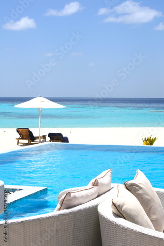 Beautiful swimming pool with a tropical beach with a deckchairs on a white sand beach with turquoise blue water sea in background,  in the turquoise of the laggon in an atoll of Maldives Islands.